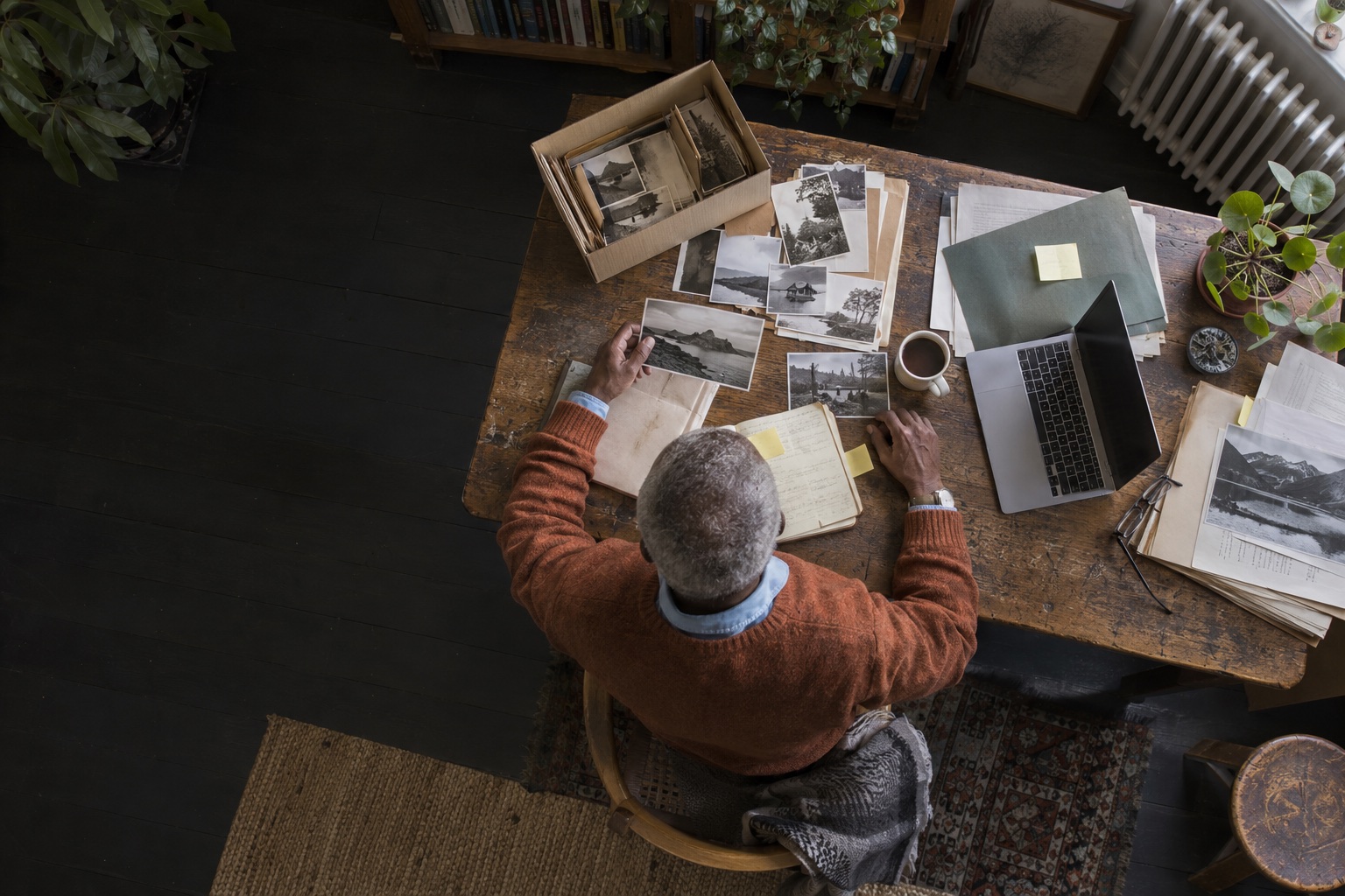 Directly overhead view of an older man in a rust cardigan sorting archive photographs and working papers in a home office.